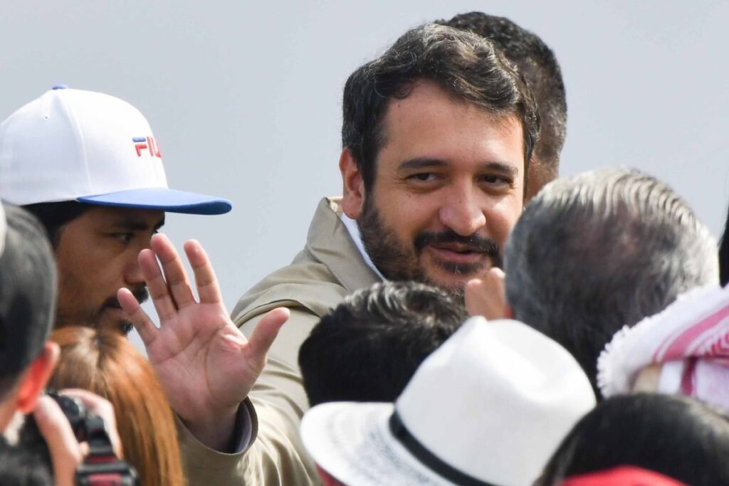 Andrés Manuel López Beltrán, bearded and wearing a beige jacket, smiles and waves from inside a crowd at a Morena event.