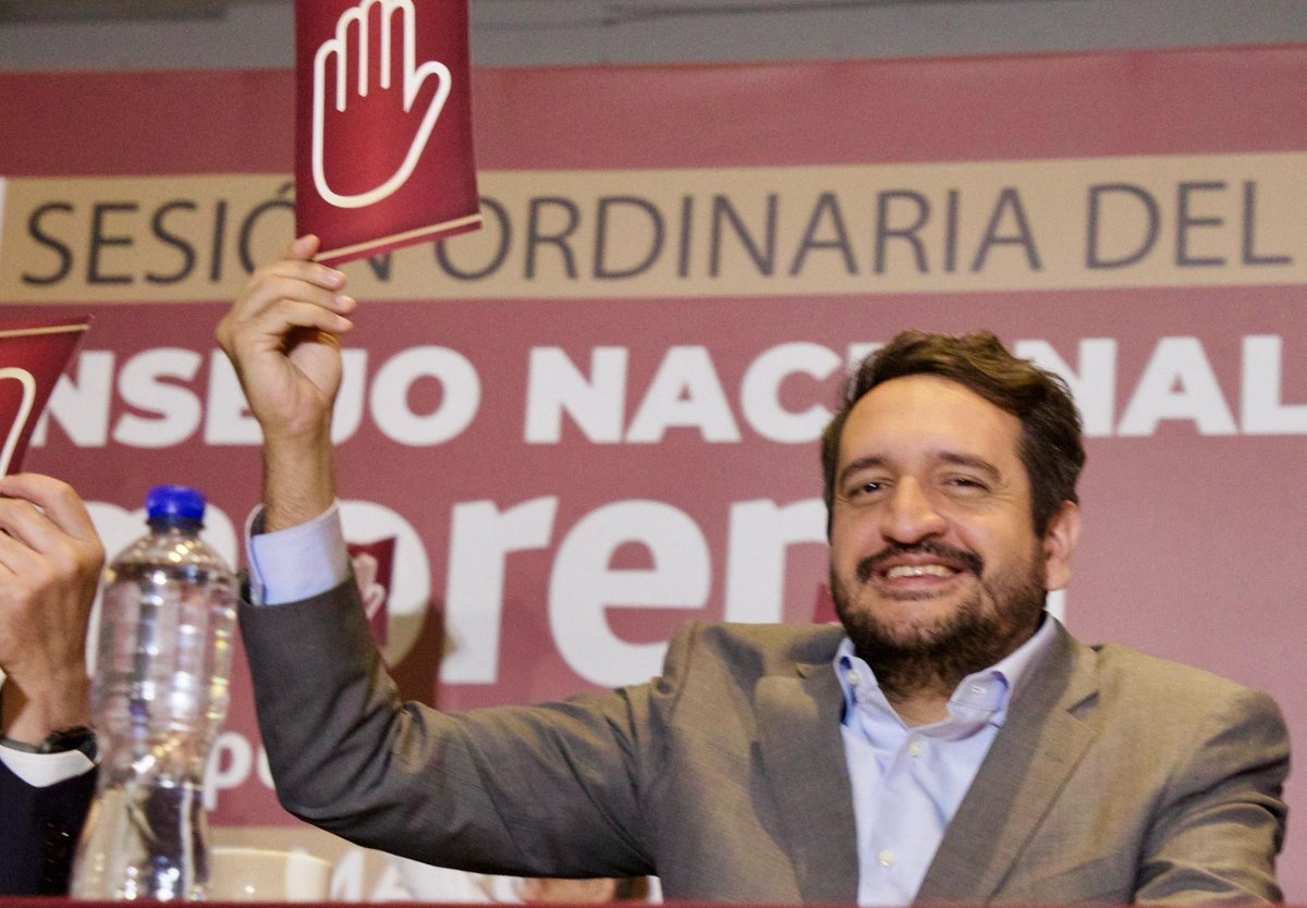 Andrés Manuel López Beltrán, in a gray jacket, smiles and raises a red voting card at a Morena National Council session.