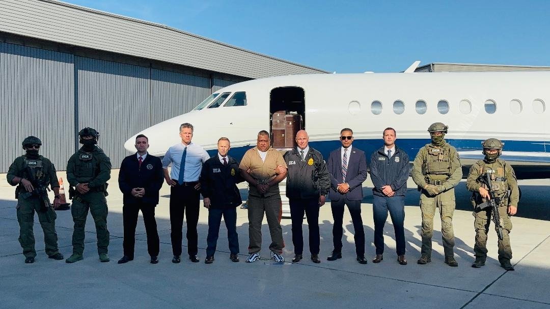 Geovany Andrés Rojas, alias Araña, in a tan uniform and shackles, standing in a line of DEA, FBI and HSI agents and U.S. officials in front of a private jet