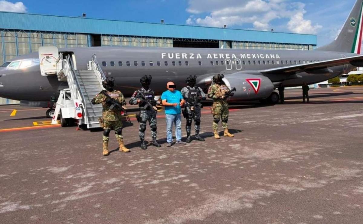 A detained man in a blue shirt, his eyes blurred, stands between four armed soldiers and National Guard members in front of a Fuerza Aérea Mexicana plane