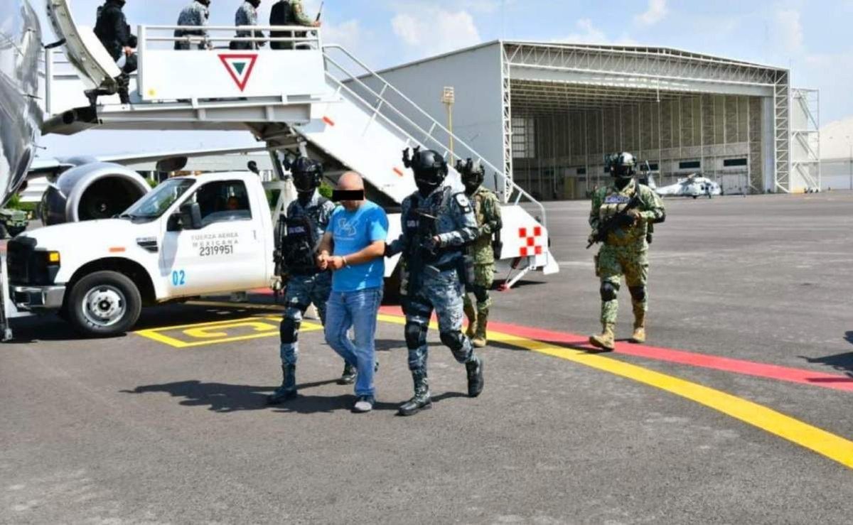 Masked National Guard and army personnel escort a detained man in a blue shirt, his eyes blurred, across an airport apron next to a Mexican Air Force plane