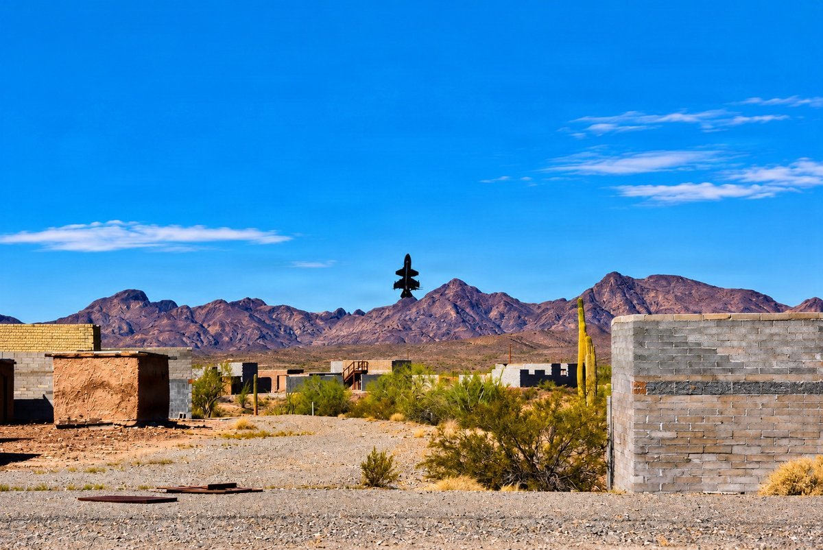 A small black interceptor drone rises above desert training buildings and saguaro cactus with purple mountains behind at Yuma Proving Ground, Arizona