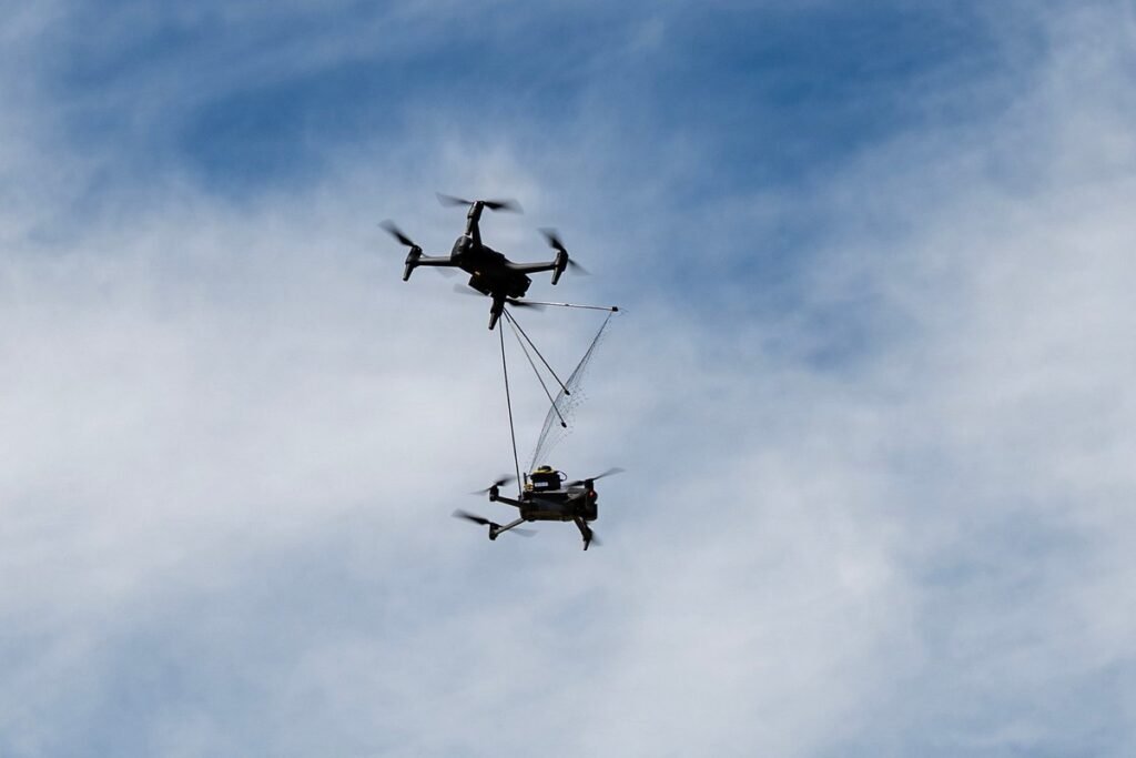 A black quadcopter trailing a net captures a second, gray "aggressor" drone in midair against a cloudy sky during a counter-drone test in Arizona