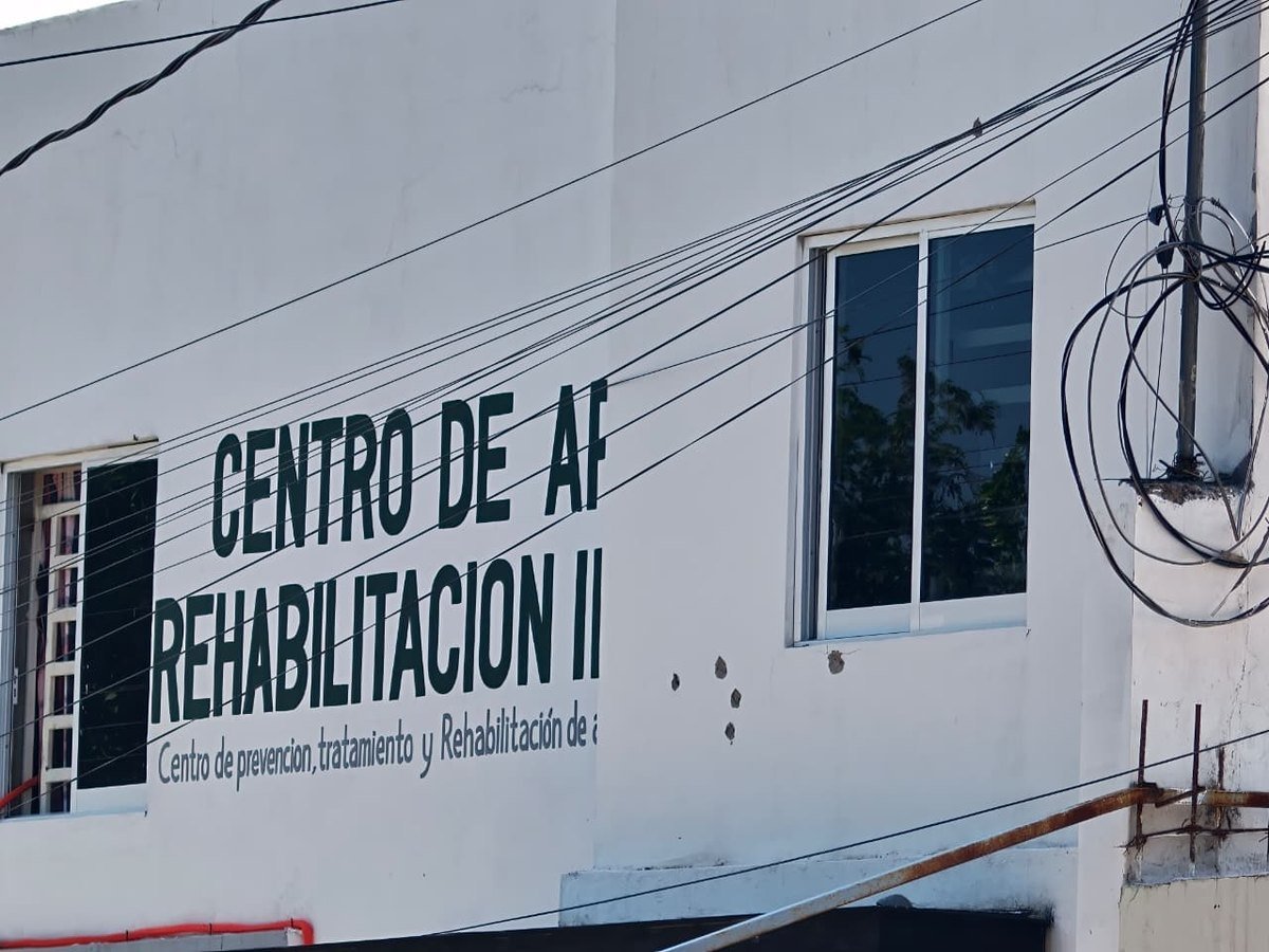 Painted sign of a drug rehabilitation center on a white wall in Culiacán, with several bullet impacts beside a window