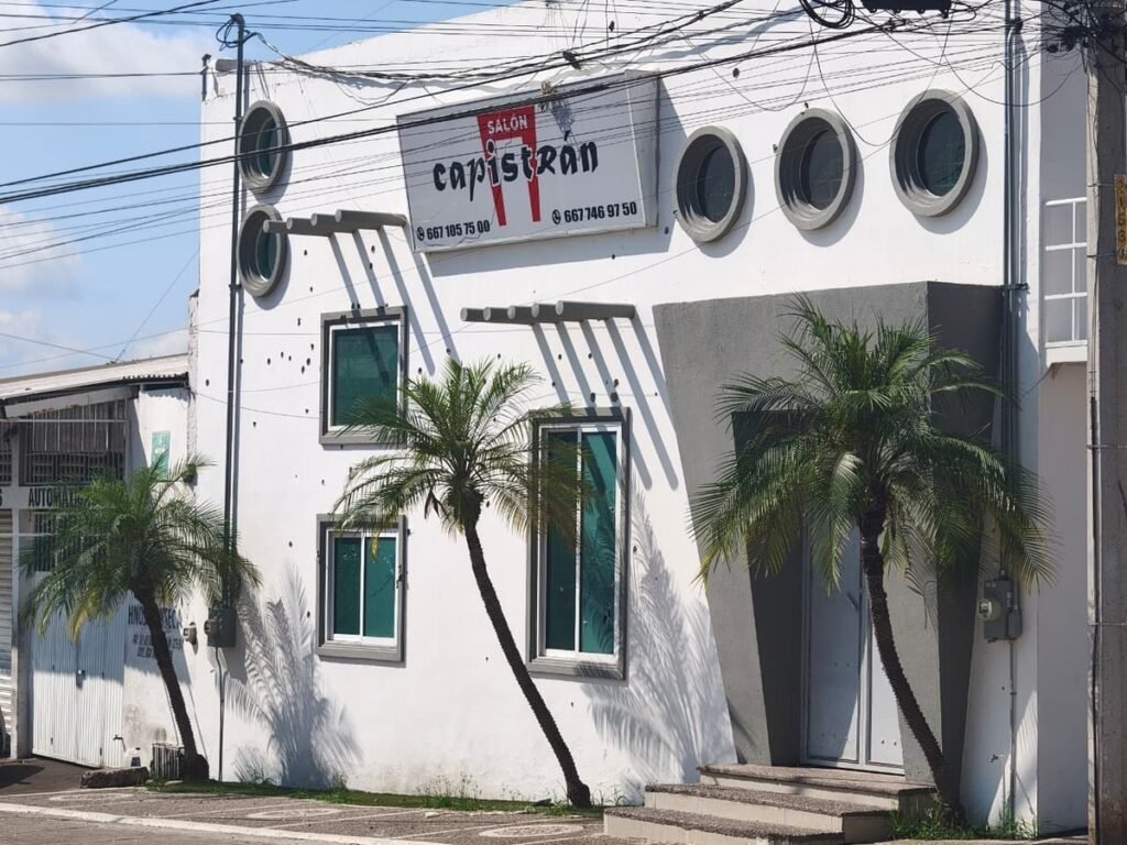 White facade of the Salón Capistrán party hall in Culiacán with bullet holes around its windows and sign, palm trees in front