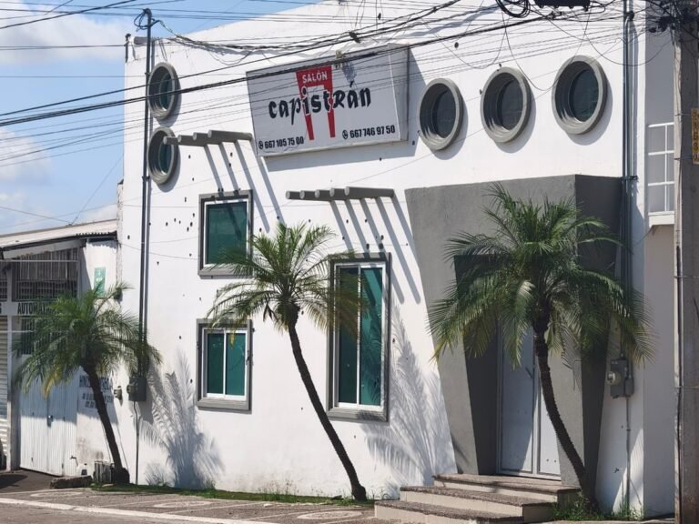 White facade of the Salón Capistrán party hall in Culiacán with bullet holes around its windows and sign, palm trees in front