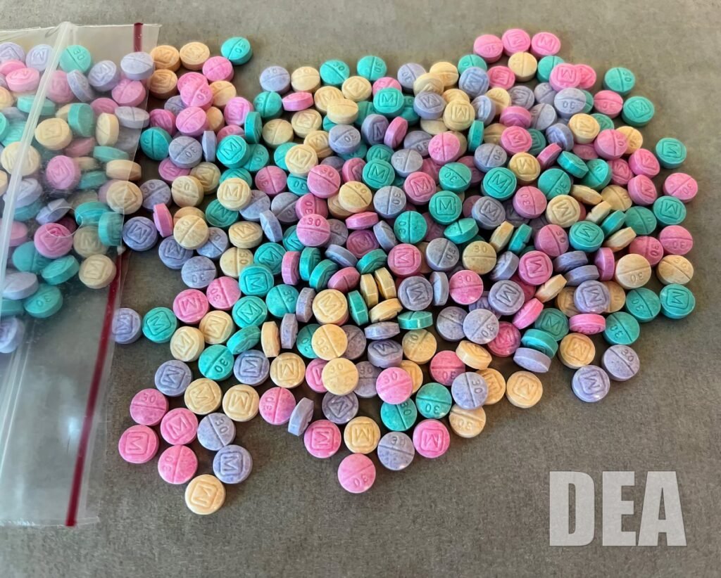 A pile of pink, purple, green, yellow and blue counterfeit fentanyl pills stamped M and 30, spilling from a plastic bag, with a DEA watermark