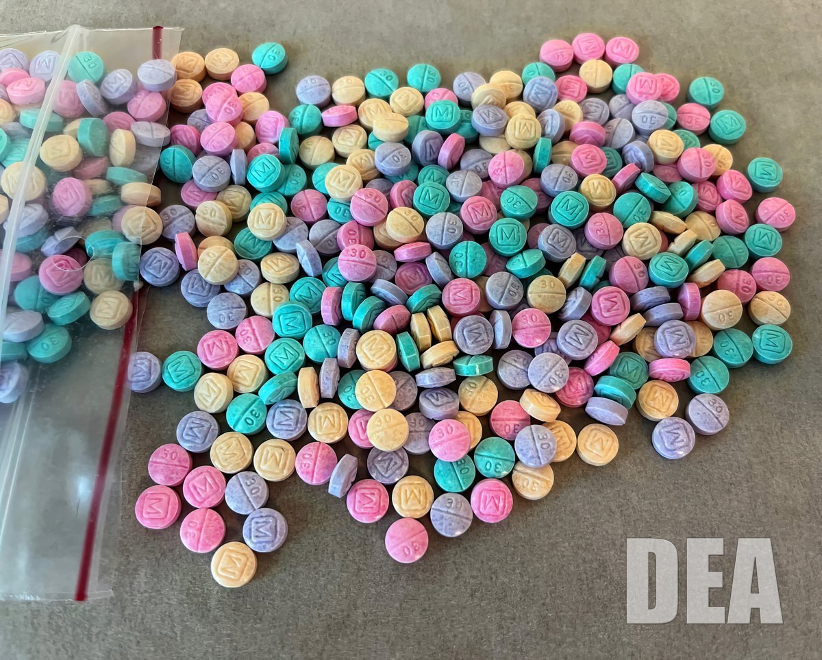 A pile of pink, purple, green, yellow and blue counterfeit fentanyl pills stamped M and 30, spilling from a plastic bag, with a DEA watermark