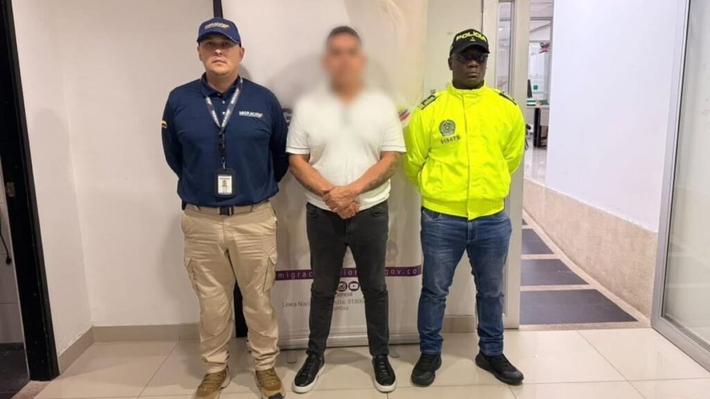A man in a white T-shirt with his face blurred stands between a Migración Colombia officer in a navy cap and a Colombian police officer in a yellow jacket in an office hallway