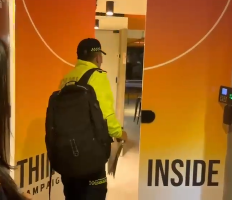 A Colombian police officer in a yellow jacket carrying a backpack walks through an orange office doorway during a search