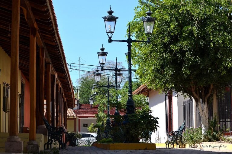 A quiet cobblestone street in El Quelite, Sinaloa, lined with a tiled portico, ornate lampposts and trees