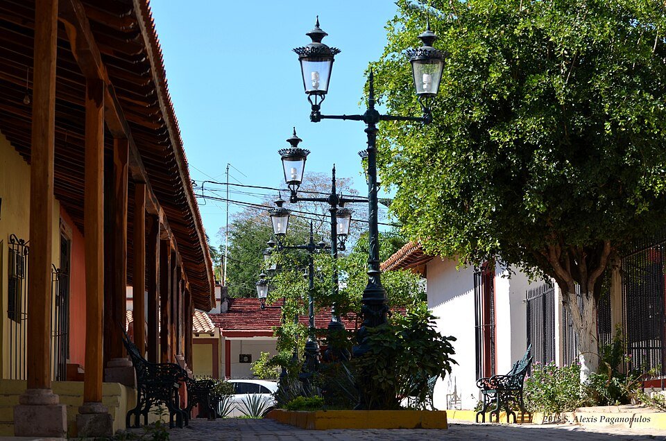 A quiet cobblestone street in El Quelite, Sinaloa, lined with a tiled portico, ornate lampposts and trees