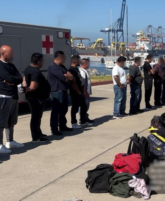 A line of adult men, faces blurred, stand beside a Navy ambulance on a dock at the port of Ensenada, with bags on the ground and cranes and ships behind them