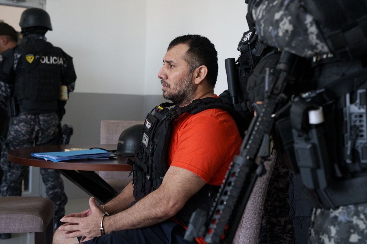 José Adolfo Macías Villamar, alias Fito, sits handcuffed and guarded by armed Ecuadorian police