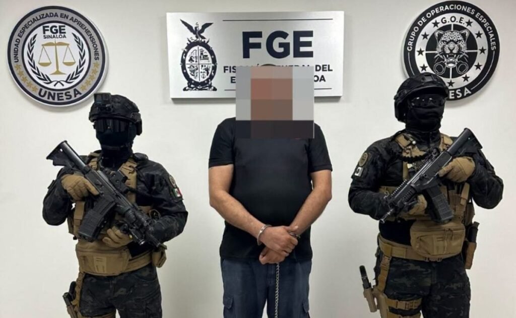 Florentino “N,” 44, stands handcuffed with his face pixelated between two armed agents in front of a Sinaloa state prosecutor's office (FGE) sign