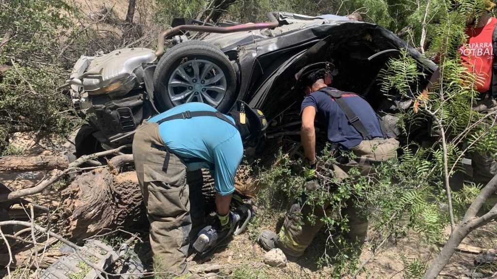 Two firefighters cut into an overturned, crushed dark sedan lying in brush and fallen tree limbs