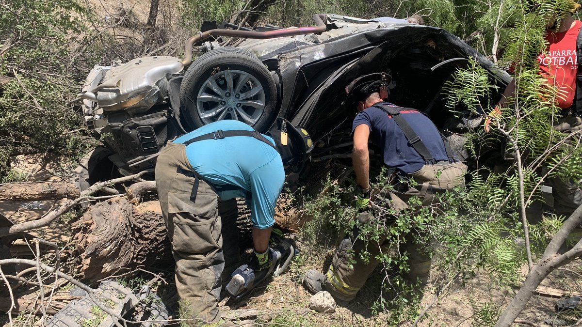 Two firefighters cut into an overturned, crushed dark sedan lying in brush and fallen tree limbs
