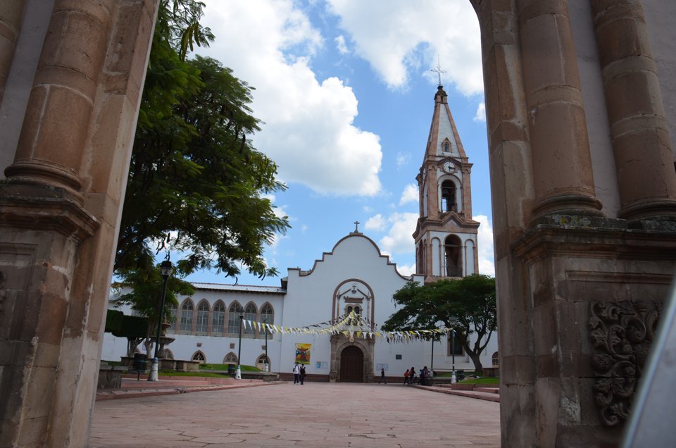 The white facade and bell tower of the Templo de San Agustín in Jacona, Michoacán, framed by stone columns under a partly cloudy sky