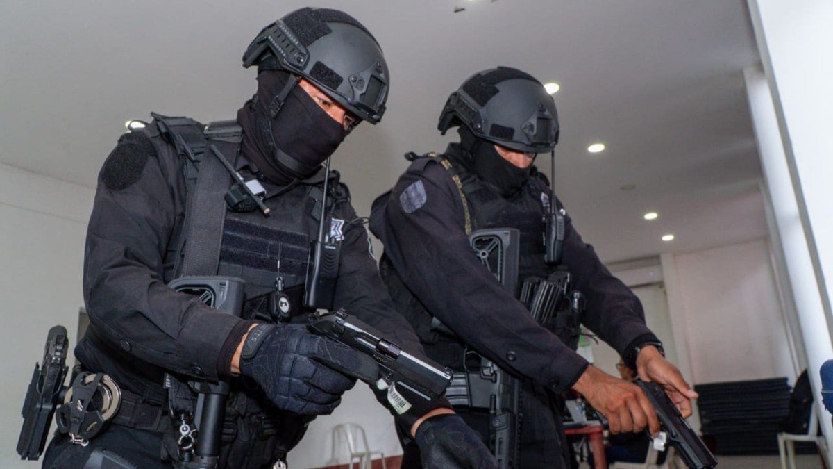 Two masked, helmeted Oaxaca state security officers in black tactical gear inspect handguns.