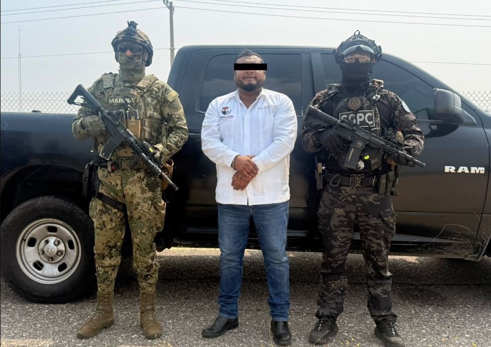 A man in a white shirt, his eyes covered by a black bar, stands between an armed Mexican Navy marine and an armed federal SSPC officer in front of a black pickup truck.