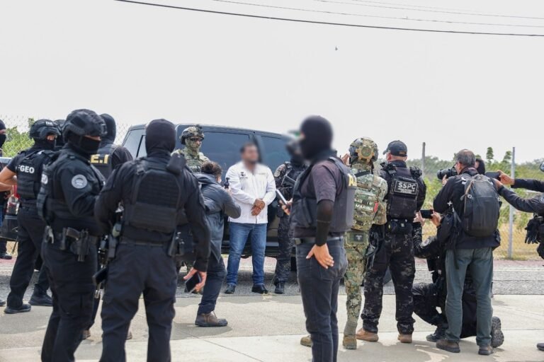 Masked Oaxaca state agents, Navy personnel and federal SSPC officers surround a detained man in a white guayabera, his face blurred, beside a black pickup as photographers take pictures.