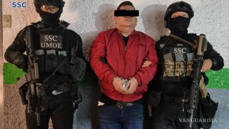 A man in a red jacket with his eyes covered by a black bar stands handcuffed between two masked, armed Mexico City police officers wearing SSC UMOE vests
