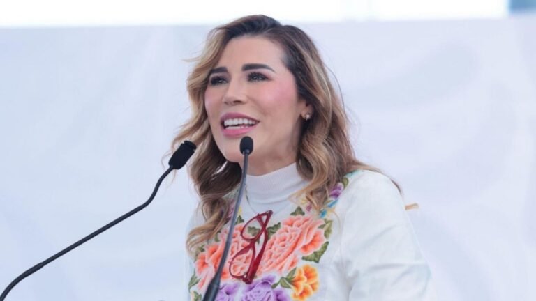 Baja California Gov. Marina del Pilar Ávila Olmeda speaking at a podium with two microphones, wearing a white blouse with embroidered flowers