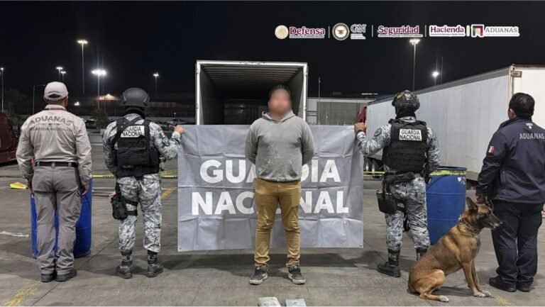 A detained truck driver with his face blurred stands in front of a Guardia Nacional banner and an open trailer at night, flanked by National Guard troops, customs officers and a sniffer dog
