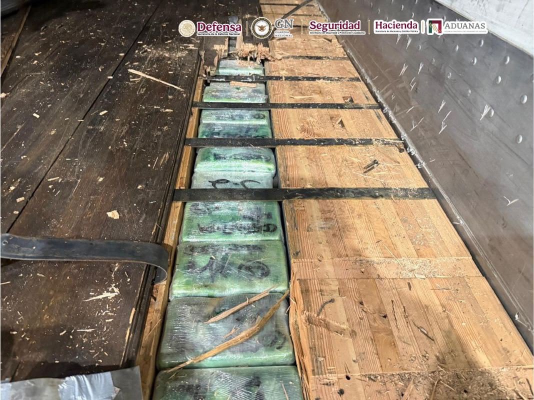 Green plastic-wrapped packages marked with black writing lie in a narrow compartment cut into the wooden floor of a trailer