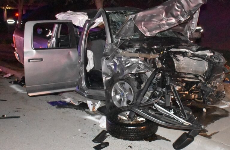 The wrecked front end of a silver Ram pickup at night, its hood crumpled and a front wheel torn loose, with debris on the pavement
