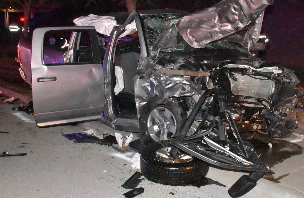 The wrecked front end of a silver Ram pickup at night, its hood crumpled and a front wheel torn loose, with debris on the pavement