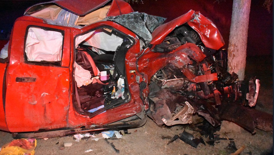A red pickup truck crushed against a tree at night, its cab and front end mangled
