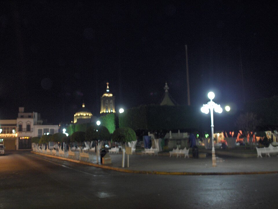 The main plaza of Puruándiro, Michoacán, at night, with a lit church dome and tower behind trimmed trees