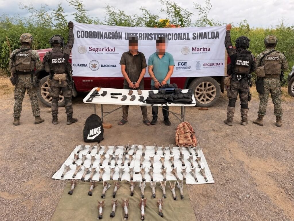 Two handcuffed men with blurred faces stand behind a table of seized rifles, magazines and tactical vests, with rows of finned drone-launched explosives laid out on tarps in front and soldiers and state police on either side