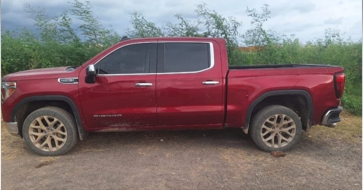 A red GMC Sierra crew-cab pickup truck without plates parked on a dirt lot in front of green brush