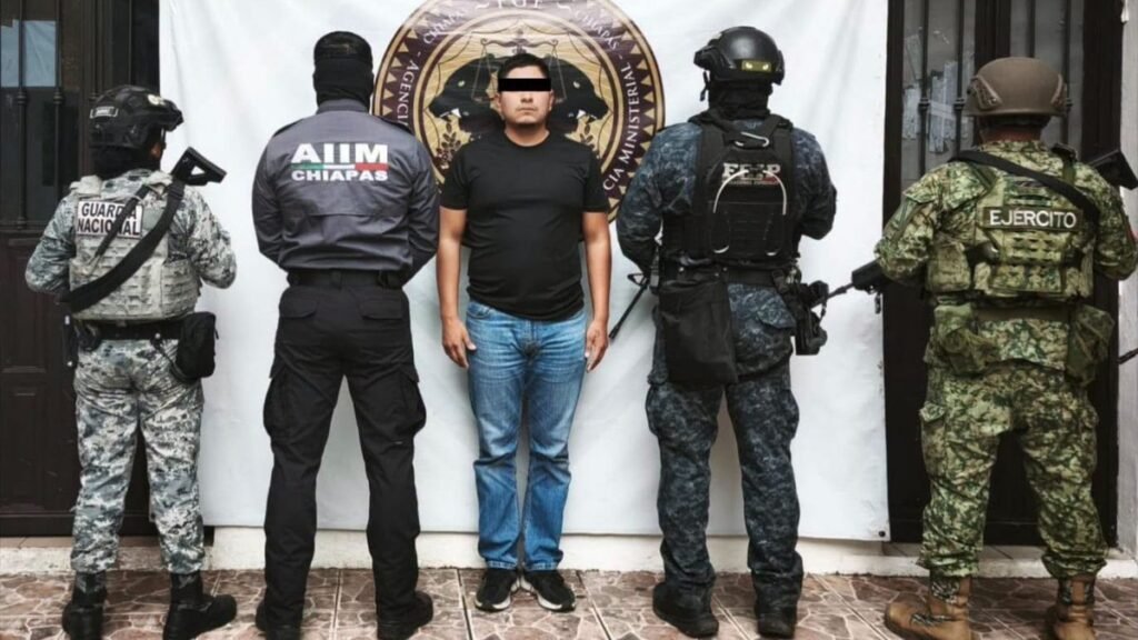 A man in a black T-shirt and jeans, eyes blacked out, stands between masked National Guard, Chiapas state investigative police and Army personnel in front of a Chiapas Attorney General's Office banner