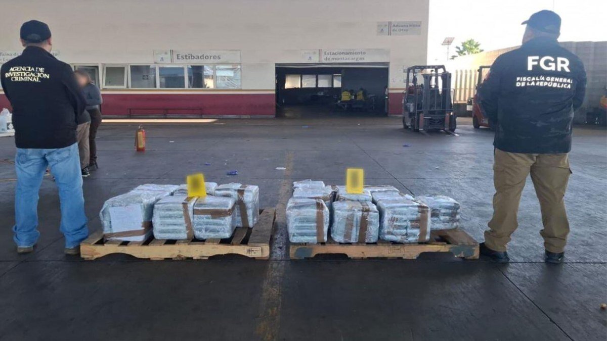 Two pallets of plastic-wrapped packages on the pavement of the Otay customs yard in Tijuana, watched by an agent in an Agencia de Investigación Criminal jacket and another in an FGR jacket