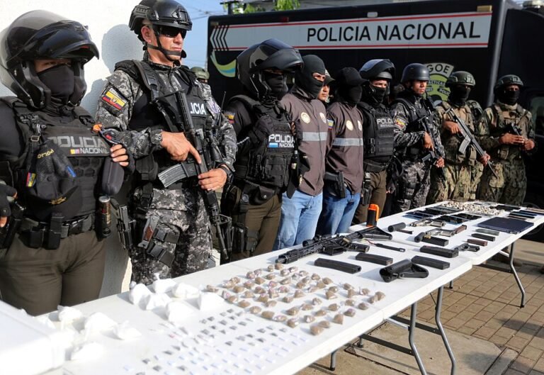 Armed Ecuadorian National Police officers in helmets and tactical gear stand behind tables of seized rifles, pistols, magazines and packaged drugs in front of a Policía Nacional truck