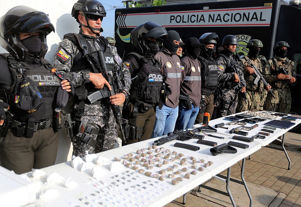 Armed Ecuadorian National Police officers in helmets and tactical gear stand behind tables of seized rifles, pistols, magazines and packaged drugs in front of a Policía Nacional truck
