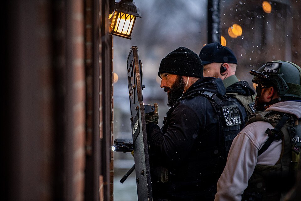 Deputy U.S. Marshals in fugitive task force vests stand with a ballistic shield outside a brick building during an enforcement operation
