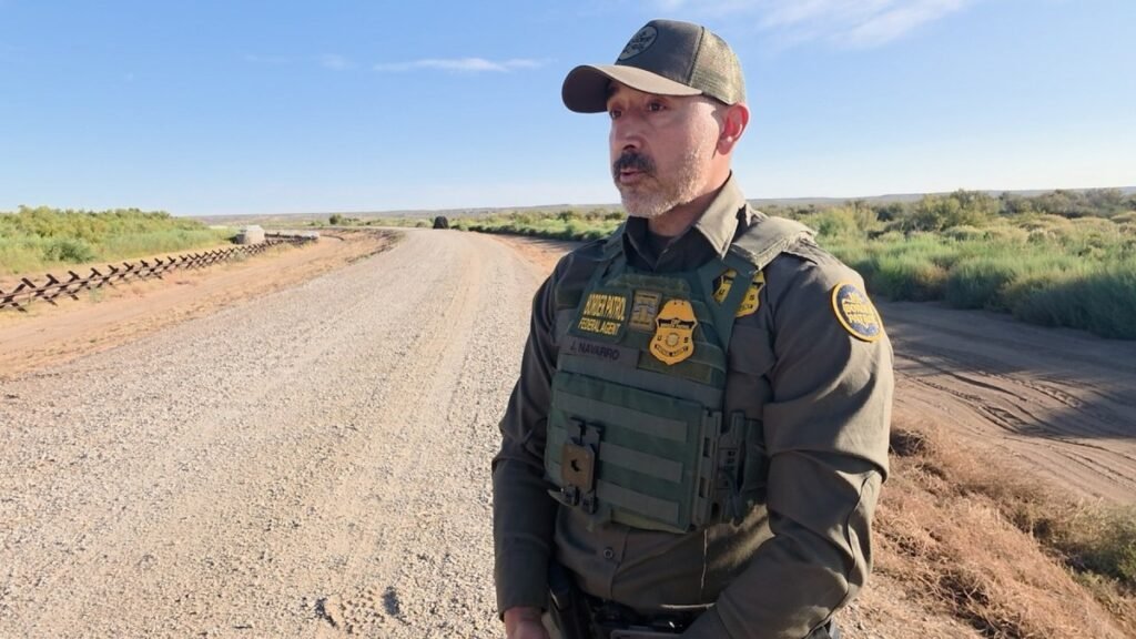 A U.S. Border Patrol agent in a cap and tactical vest stands on a gravel road beside low steel vehicle barriers and green brush in rural West Texas