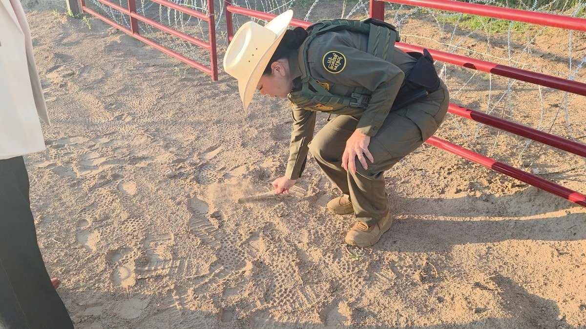 A Border Patrol agent in a straw hat crouches by a red metal gate and concertina wire, sweeping a branch across footprints in the sand