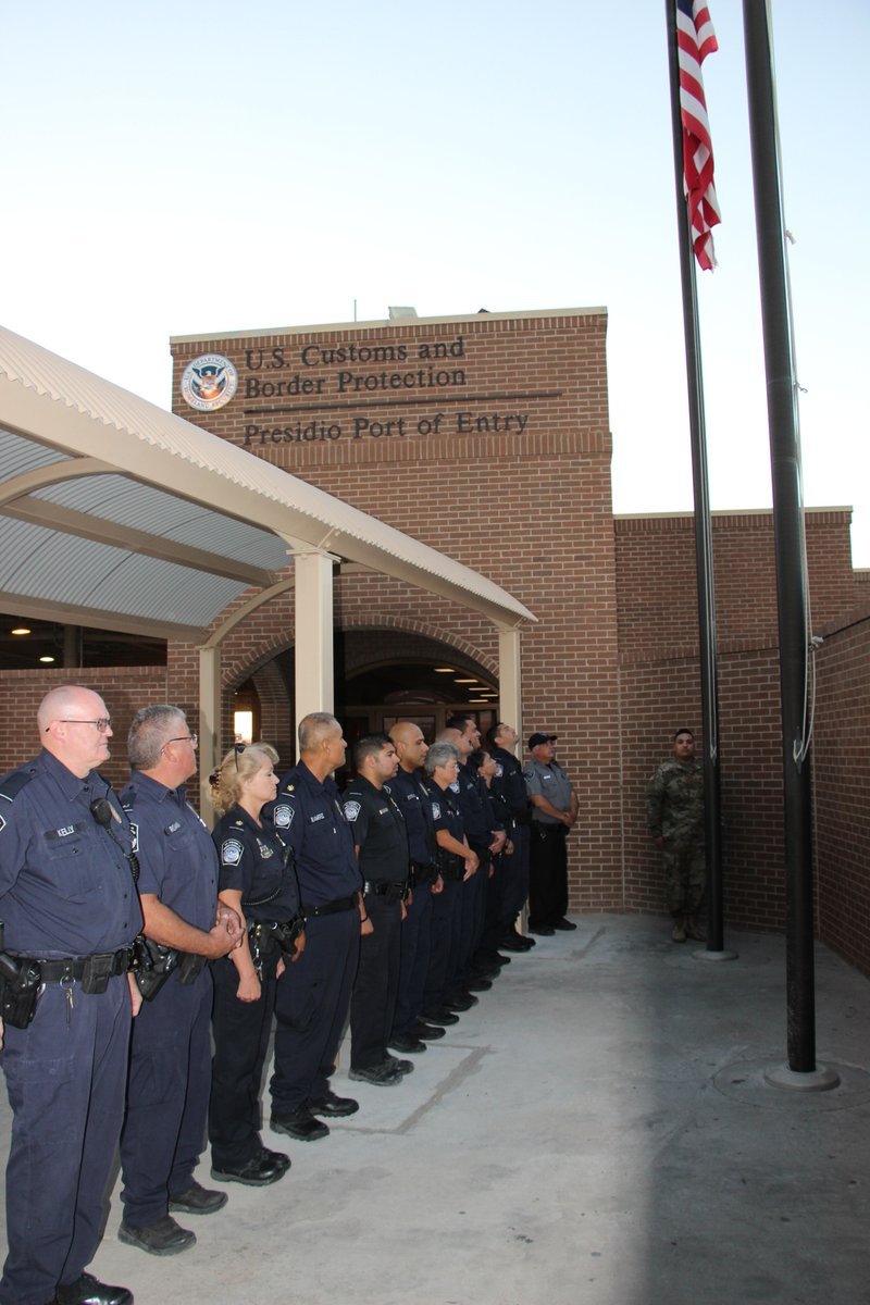 Uniformed CBP officers stand in a line outside the brick U.S. Customs and Border Protection Presidio Port of Entry building beside a U.S. flag