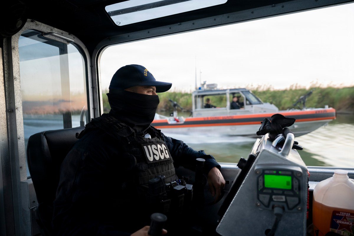 Masked Coast Guard crew member in a USCG vest at the helm of a boat as an orange-striped Coast Guard response boat passes on the Rio Grande