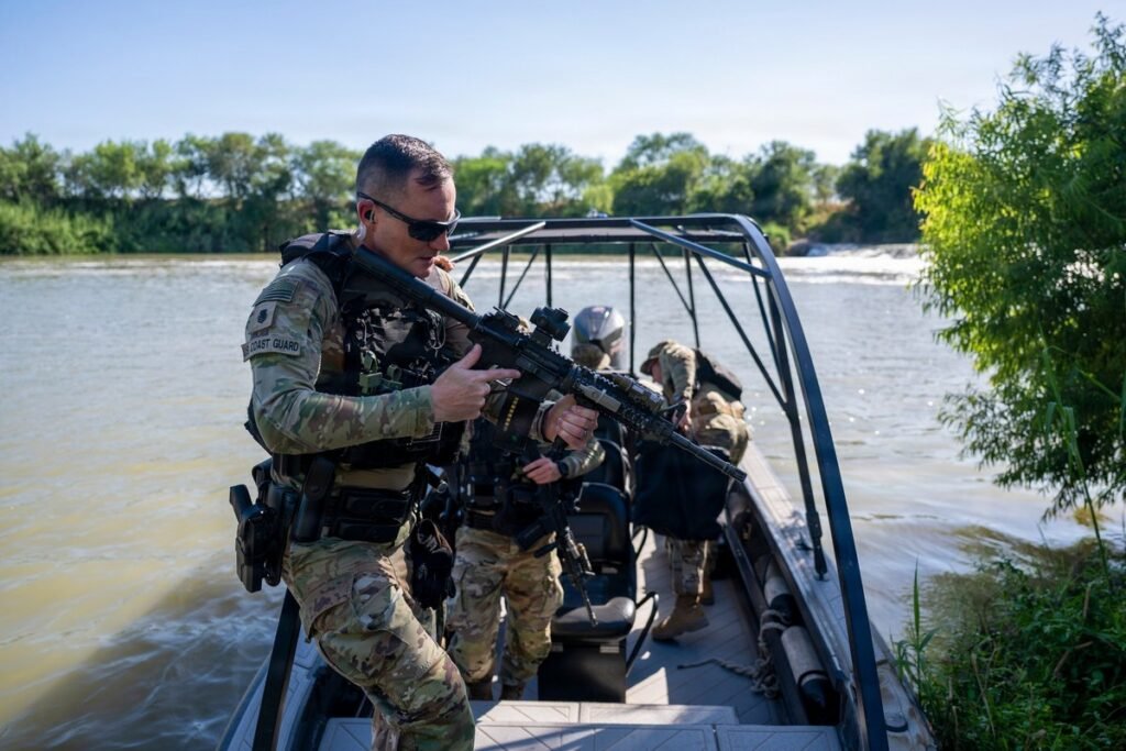 Armed U.S. Coast Guard member in camouflage and body armor steps off a small patrol boat at the bank of the Rio Grande, with other crew members aboard