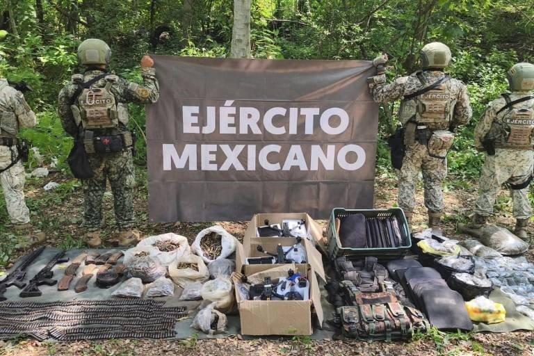 Mexican Army soldiers hold an 'Ejército Mexicano' banner behind seized rifles, ammunition belts, cartridges, drones in boxes and tactical vests laid out in a forest clearing