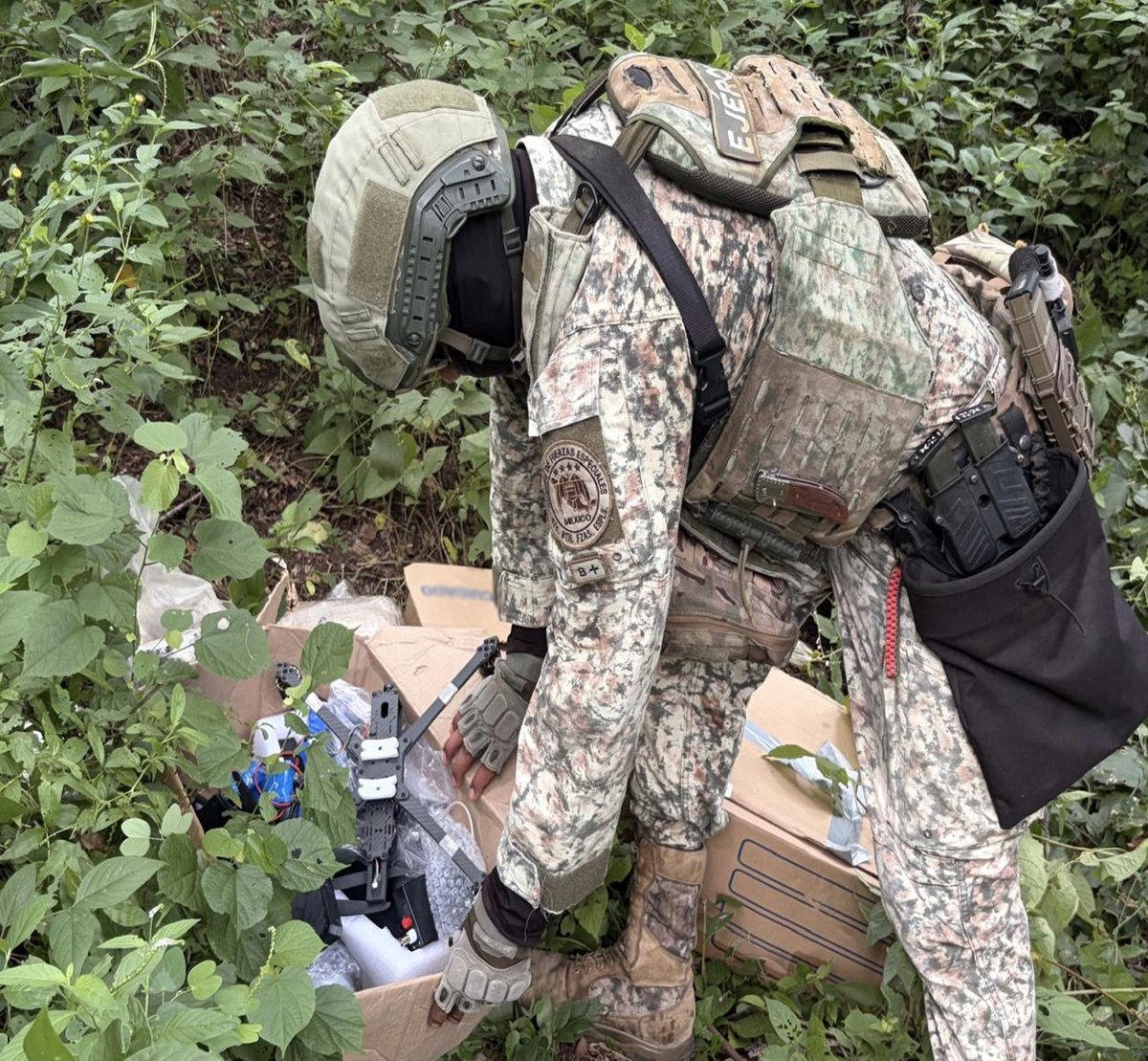 Mexican Army special forces soldier bends over a cardboard box holding a carbon-frame drone in brush in Concordia, Sinaloa