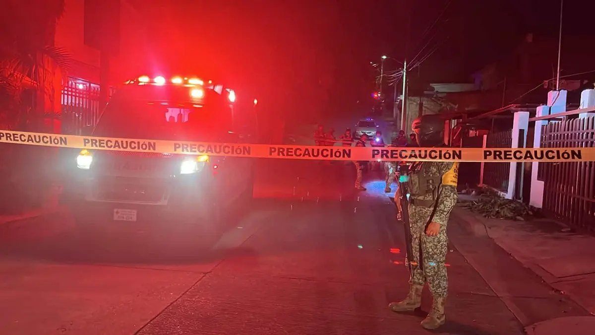 An ambulance with flashing lights and a soldier behind yellow caution tape on a residential street in Culiacán at night.