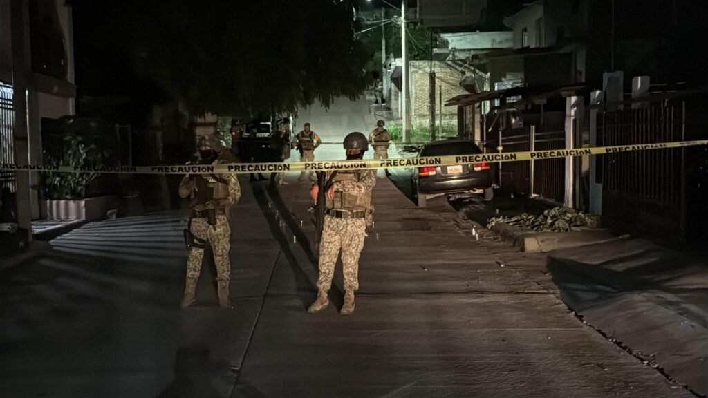 Mexican Army soldiers in camouflage stand guard behind yellow 'Precaución' tape on a dark residential street in Culiacán's 10 de Mayo neighborhood at night.