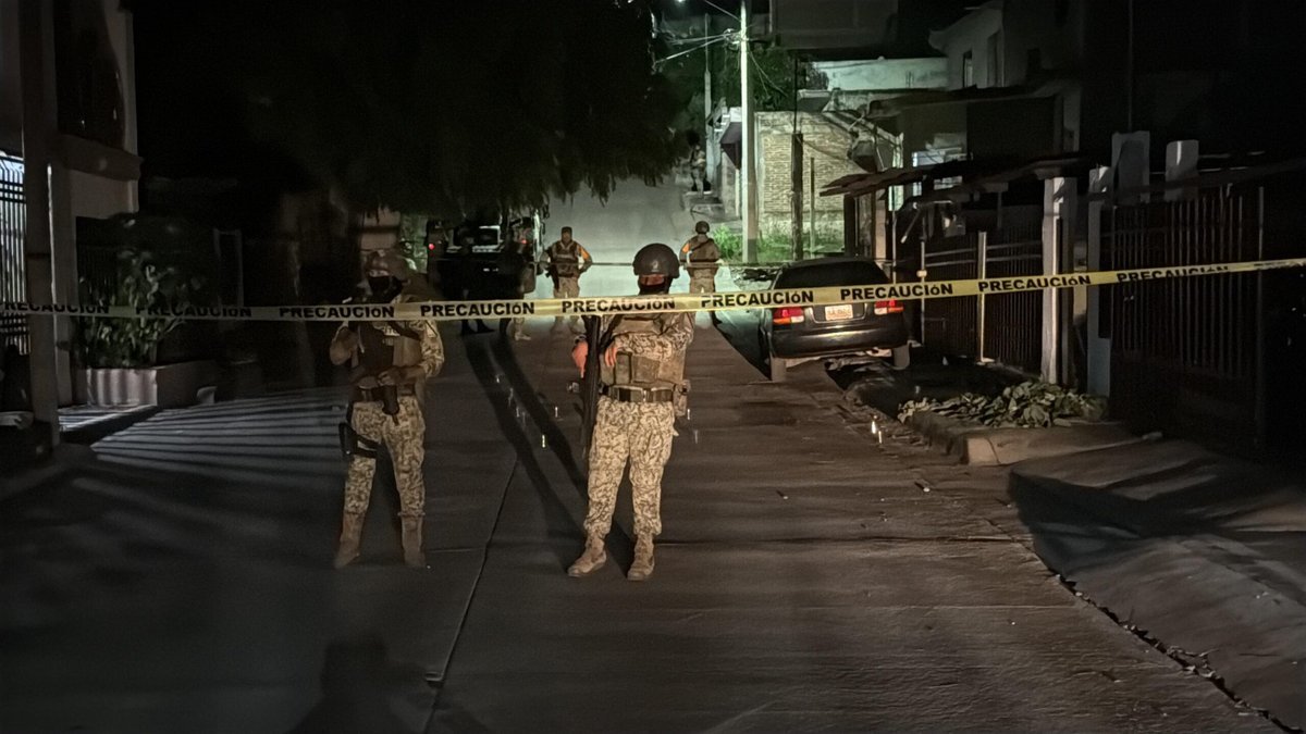 Mexican Army soldiers in camouflage stand guard behind yellow 'Precaución' tape on a dark residential street in Culiacán's 10 de Mayo neighborhood at night.